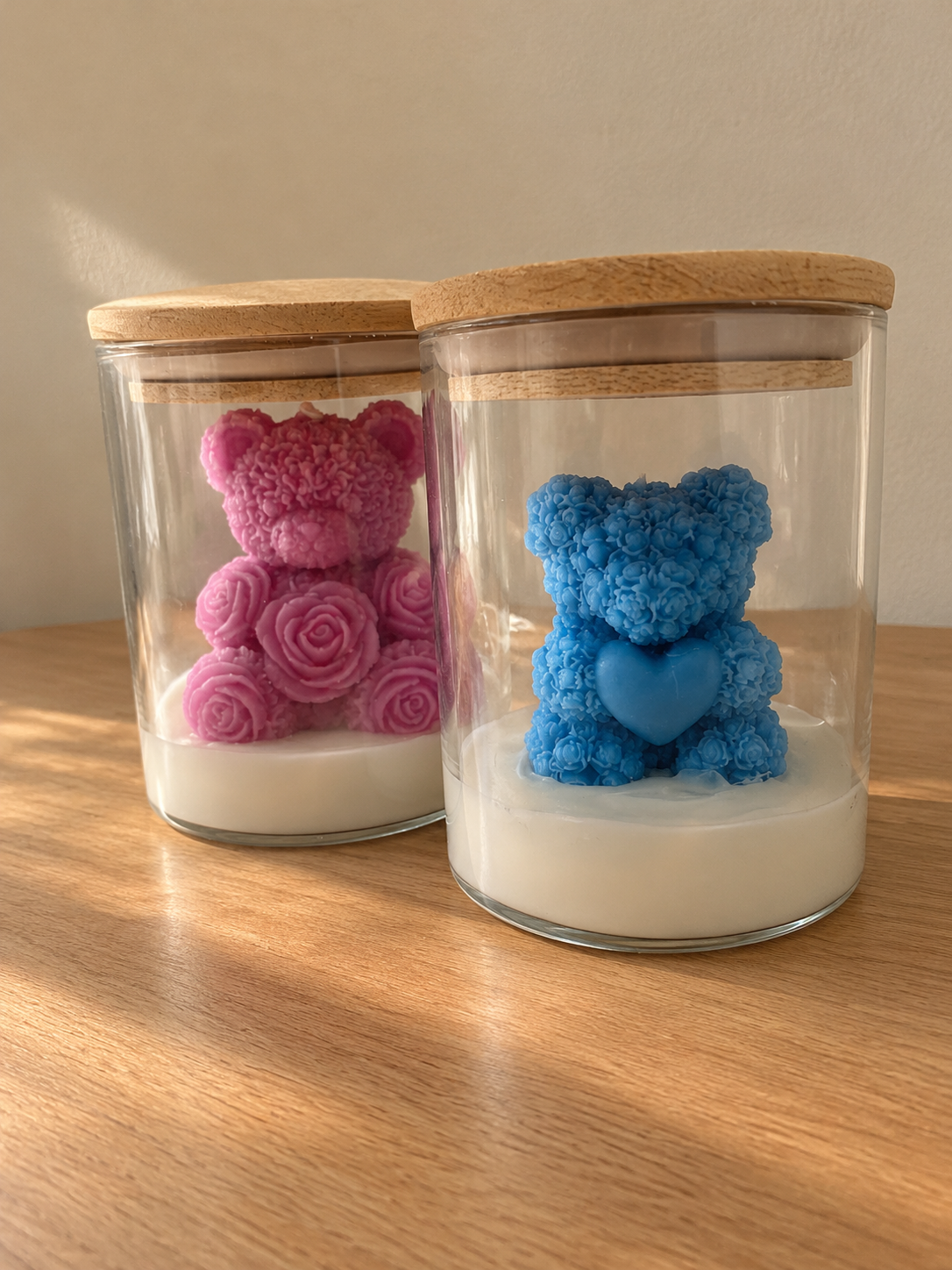 Two glass jars with wooden lids, each holding a rose-textured bear candle, one pink and one blue