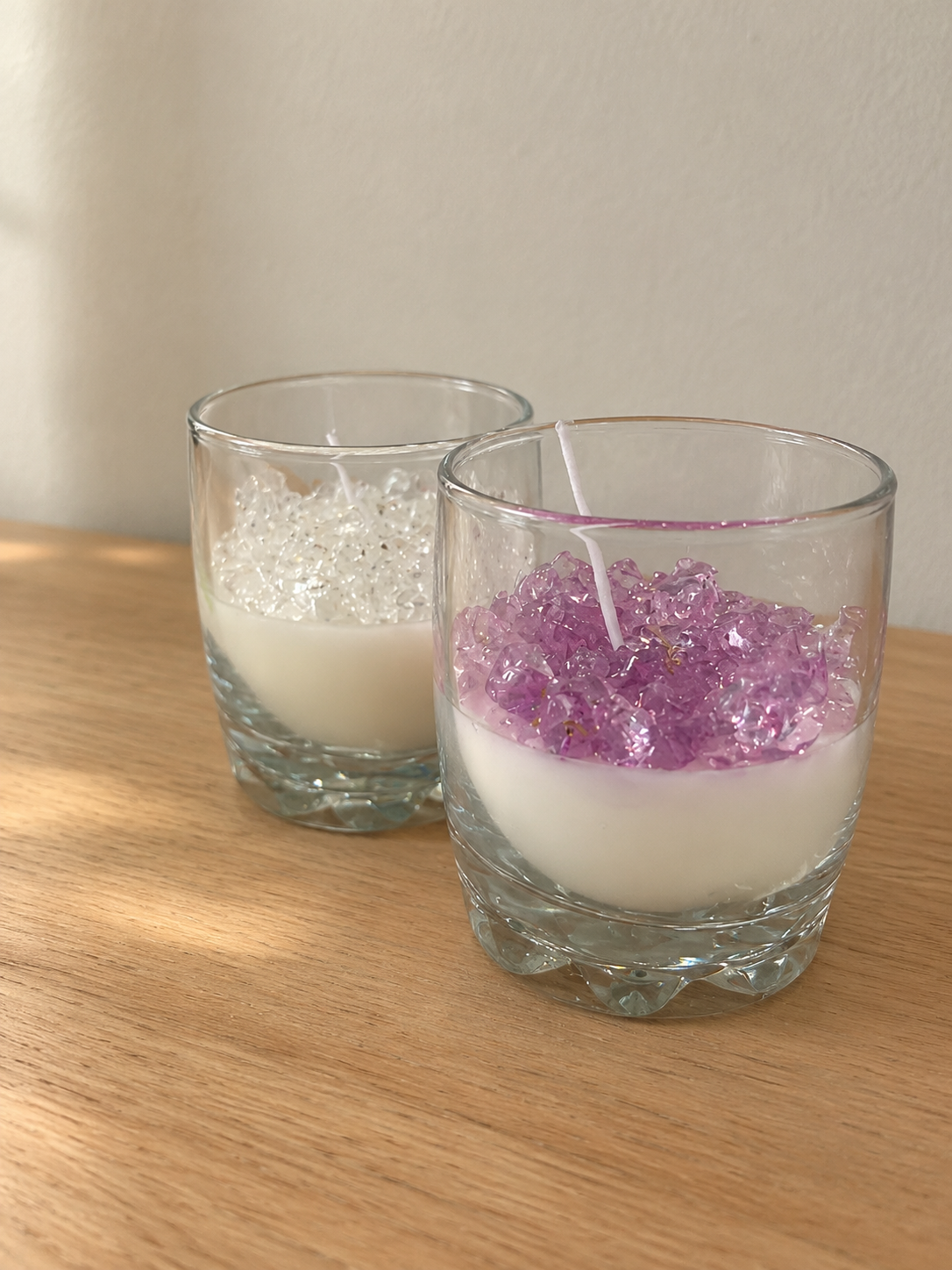 Two glass candles of white wax topped with clear and lilac crystal-like pieces